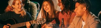 Gruppo di amici che ride, canta e suona strumenti musicali intorno a un falò serale, davanti a una tenda.