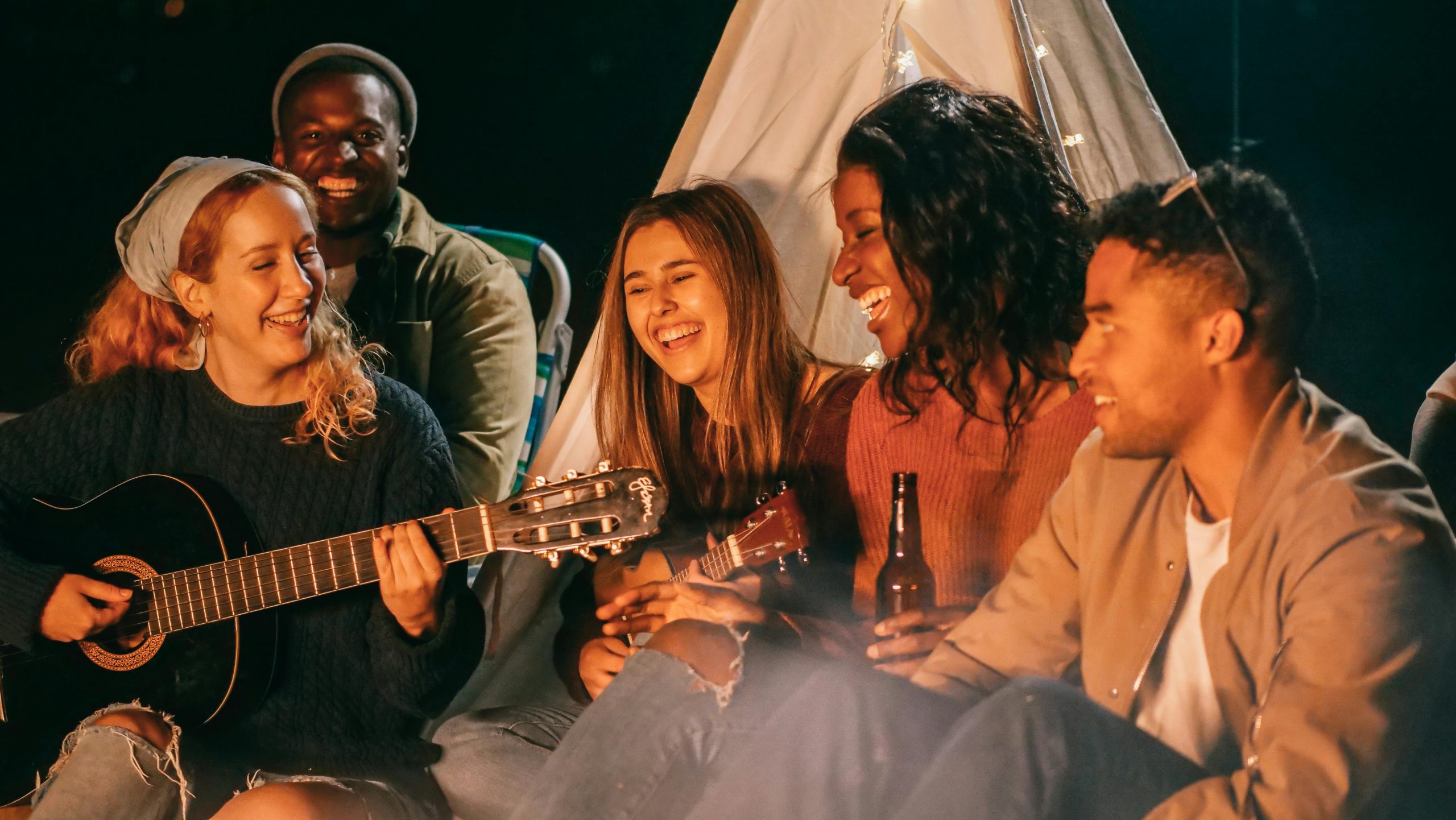 Gruppo di amici che ride, canta e suona strumenti musicali intorno a un falò serale, davanti a una tenda.